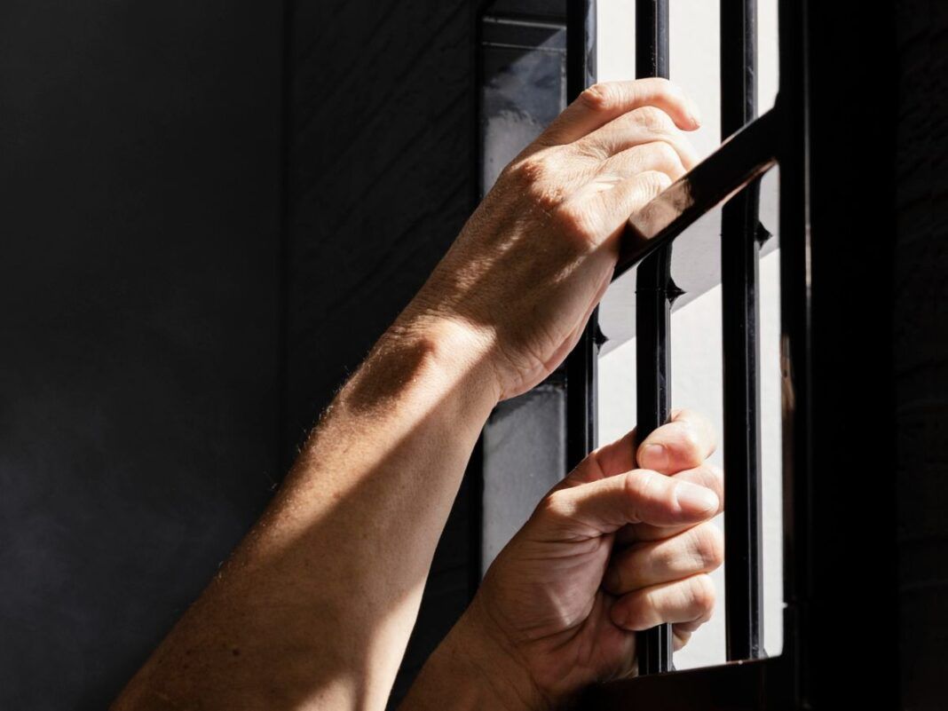 Imagem mostra as mãos de um detento segurando as grades da prisão