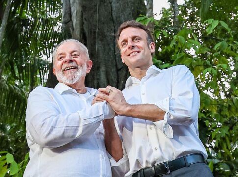 A imagem, com uma iluminação natural e suave, apresenta o Presidente do Brasil, Luiz Inácio Lula da Silva, e o Presidente da França, Emmanuel Macron, de pé, lado a lado, em um ambiente que parece ser uma floresta, possivelmente a Amazônia, como indicam os grandes troncos de árvores e a folhagem densa e escura no fundo.