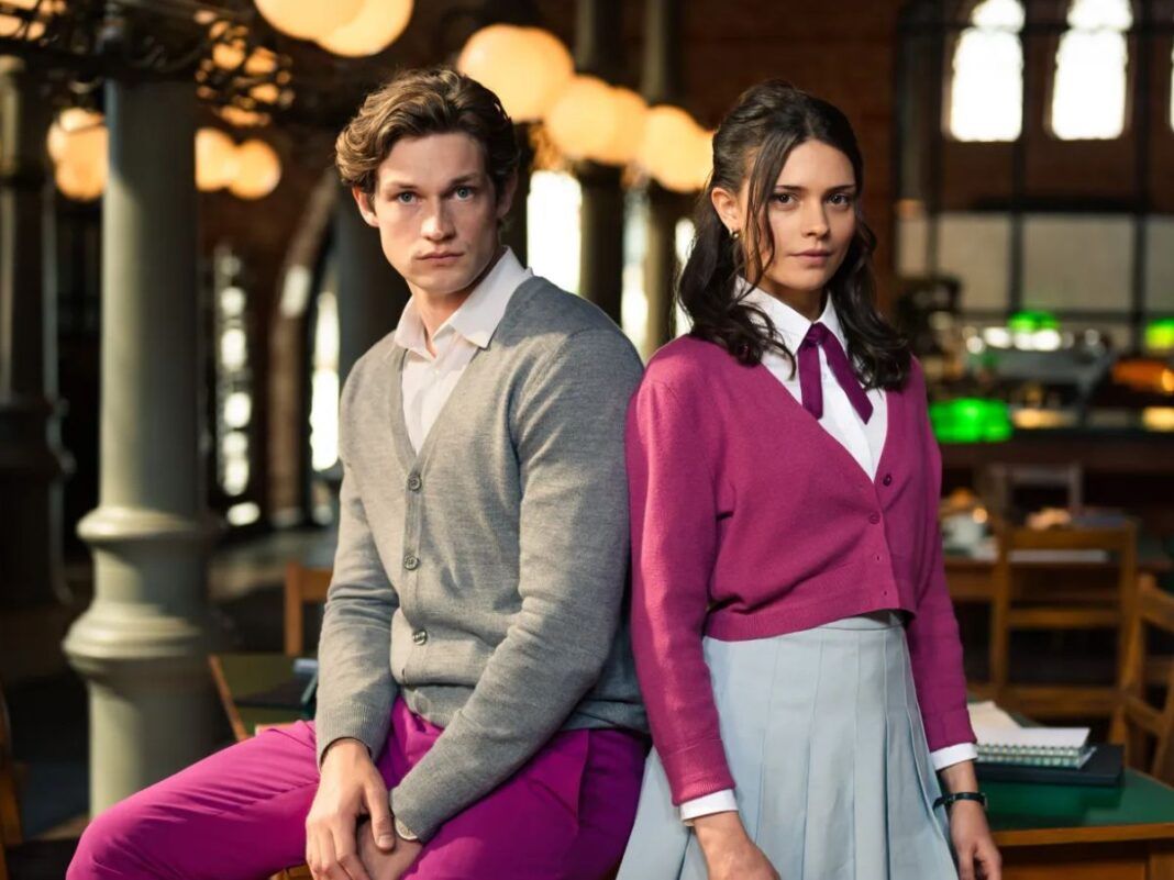 Na foto há um homem loiro sentado em uma mesa e do lado há uma mulher de pé. Ambos estão uma biblioteca.