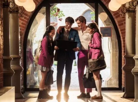 Na foto estão 4 jovens com uniformes que possuem cores cinza r osa. Eles estão em um corredor e olhando para um tablet.