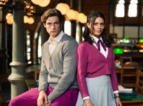 Na foto há um homem loiro sentado em uma mesa e do lado há uma mulher de pé. Ambos estão uma biblioteca.