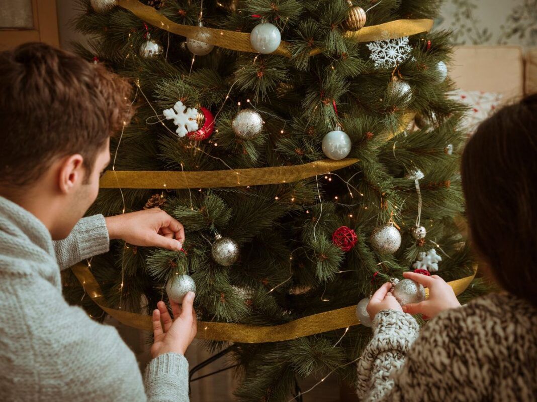 Casal colocando enfeites cinzas em uma grande árvore de Natal verde