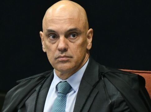 A foto é um retrato em plano médio-fechado, focado no Ministro do Supremo Tribunal Federal (STF), Alexandre de Moraes. O ministro aparece em uma pose formal, olhando diretamente para a frente ou ligeiramente para o lado direito do observador. Ele tem a cabeça raspada/calva e as sobrancelhas arqueadas, com uma expressão séria, tensa e de intensa concentração, que pode ser interpretada como desconfiança ou atenção plena. Seus olhos estão arregalados e fixos.