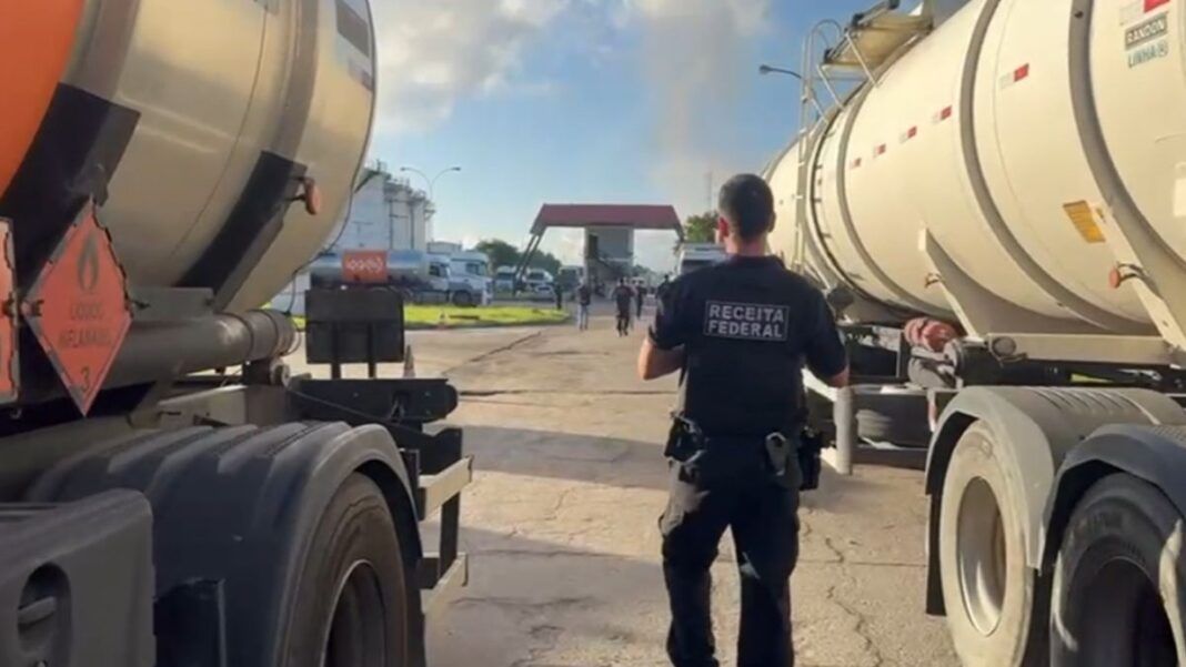 Esta é uma fotografia em plano médio, tirada ao nível do chão, em um ambiente industrial ou de pátio de caminhões sob luz natural intensa. O foco está em um agente de segurança em meio a dois caminhões-tanque.