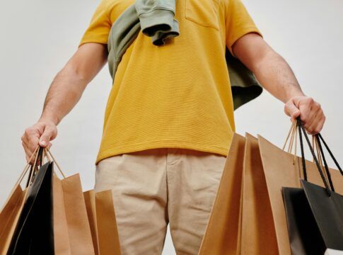 Pessoa de camiseta amarela segura várias sacolas de compras nas duas mãos. A imagem é vista de baixo, destacando braços, roupas e as sacolas marrons e pretas.