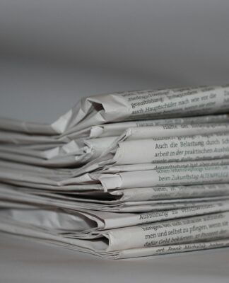 A imagem mostra uma pilha de jornais dobrados, com o papel ligeiramente amassado e textos impressos visíveis nas laterais. Tudo está em tons de cinza, com fundo neutro e desfocado.