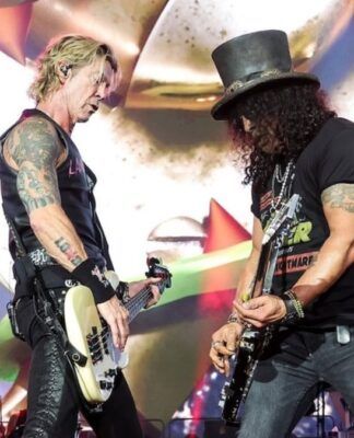 Esta é uma fotografia vibrante tirada no palco durante um show da banda de rock Guns N' Roses. O foco está em dois membros da banda em um momento de performance intensa.