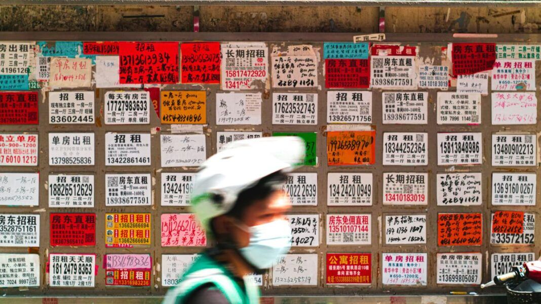 Parede coberta por dezenas de anúncios de aluguel escritos em caracteres chineses, em papéis coloridos e de tamanhos variados; na frente, desfocado, passa um ciclista usando capacete branco, máscara e colete verde.