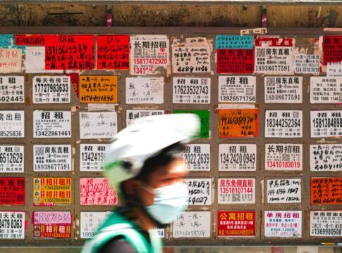 Parede coberta por dezenas de anúncios de aluguel escritos em caracteres chineses, em papéis coloridos e de tamanhos variados; na frente, desfocado, passa um ciclista usando capacete branco, máscara e colete verde.