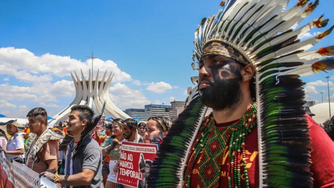 Indígenas marcham em protesto diante da Catedral de Brasília, usando cocares, pinturas corporais e segurando cartazes contra o marco temporal. A cena mostra união, resistência e defesa dos direitos dos povos originários sob um céu ensolarado.