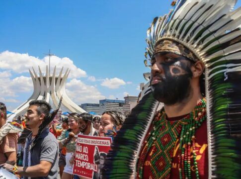 Indígenas marcham em protesto diante da Catedral de Brasília, usando cocares, pinturas corporais e segurando cartazes contra o marco temporal. A cena mostra união, resistência e defesa dos direitos dos povos originários sob um céu ensolarado.