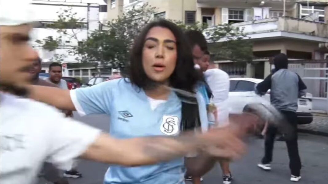 Mulher de camiseta azul-pastel do Santos surpreendida por um homem - na imagem, um borrão, pois estava em rápido movimento - de boné e vestindo camiseta branca do Santos. O homem toma o microfone da mão da repórter.
