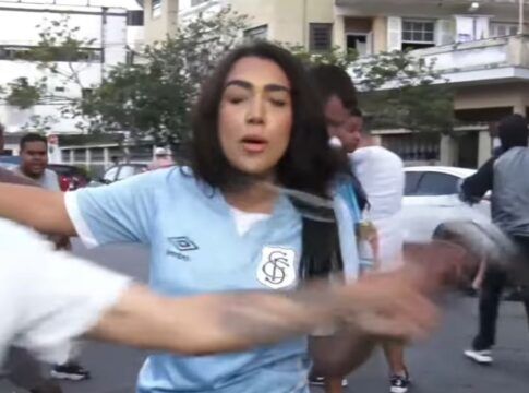 Mulher de camiseta azul-pastel do Santos surpreendida por um homem - na imagem, um borrão, pois estava em rápido movimento - de boné e vestindo camiseta branca do Santos. O homem toma o microfone da mão da repórter.