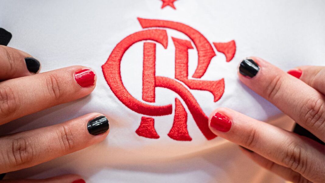 Close no escudo do Flamengo em uma camisa branca de jogo. Perto do escudo estão dedos com unhas pintadas de preto ou vermelho