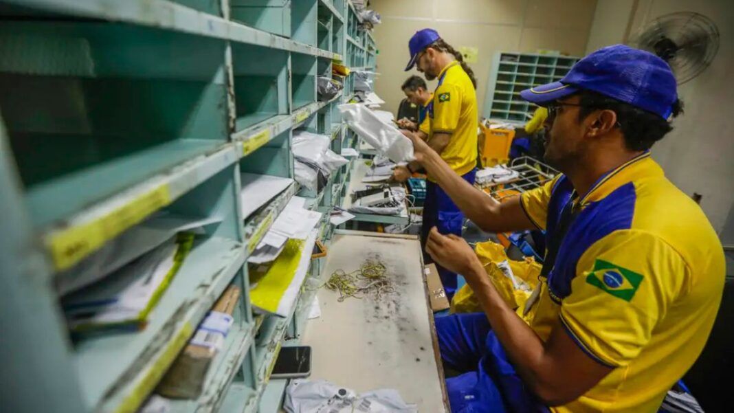 Funcionários dos Correios, uniformizados, manuseiam correspondências dentro de uma sala