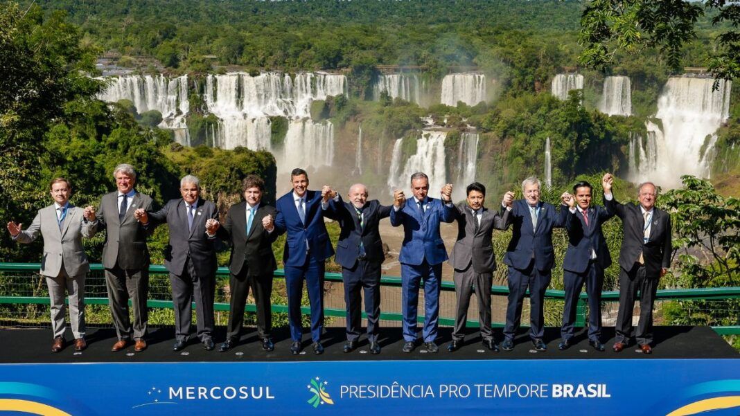 Fotografia oficial dos presidentes e chefes de delegação dos Estados Partes do Mercosul, todos em pé, de mãos dadas, à frente das Cataratas do Iguaçu