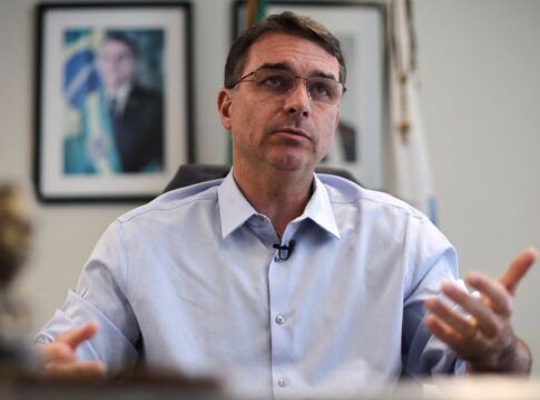 Flávio Bolsonaro gesticula durante entrevista, com foto de Jair Bolsonaro ao fundo