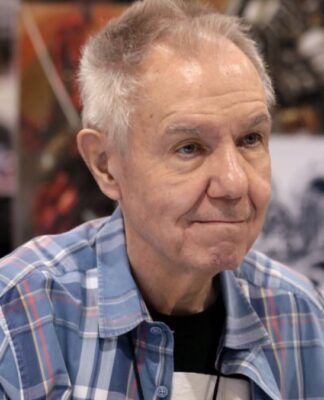 Homem mais velho, de cabelos grisalhos curtos, veste camisa xadrez azul e está sentado em um evento; ao fundo, desfocados, aparecem painéis com ilustrações coloridas e outras pessoas circulando.