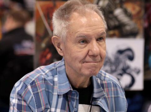 Homem mais velho, de cabelos grisalhos curtos, veste camisa xadrez azul e está sentado em um evento; ao fundo, desfocados, aparecem painéis com ilustrações coloridas e outras pessoas circulando.