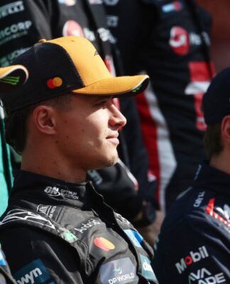 Três pilotos de automóveis sentados lado a lado, usando macacões e bonés de suas equipes de corrida. Eles olham para a frente com expressão neutra, enquanto outros membros de equipes aparecem desfocados ao fundo.