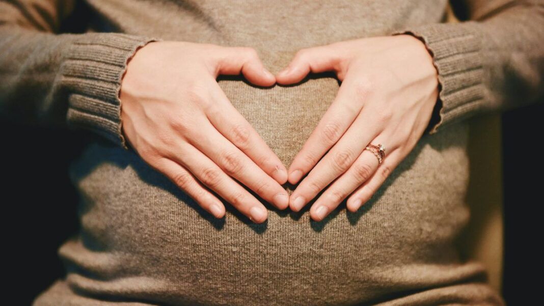 Mãos formando um coração sobre uma barriga de gestante, ambas apoiadas em um suéter de tricô bege; uma das mãos usa um anel delicado.