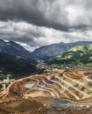 Vista panorâmica de uma grande mina a céu aberto com degraus escavados em tons de marrom e bege; no fundo, há um vale com pequenas construções e, além dele, cadeias de montanhas altas e rochosas sob um céu carregado de nuvens escuras.