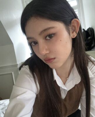 Mulher coreana de cabelos pretos, vestindo blusa branca e marrom de manga longa, tirando selfie num quarto