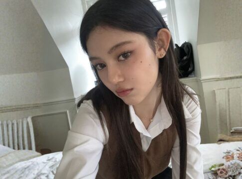 Mulher coreana de cabelos pretos, vestindo blusa branca e marrom de manga longa, tirando selfie num quarto