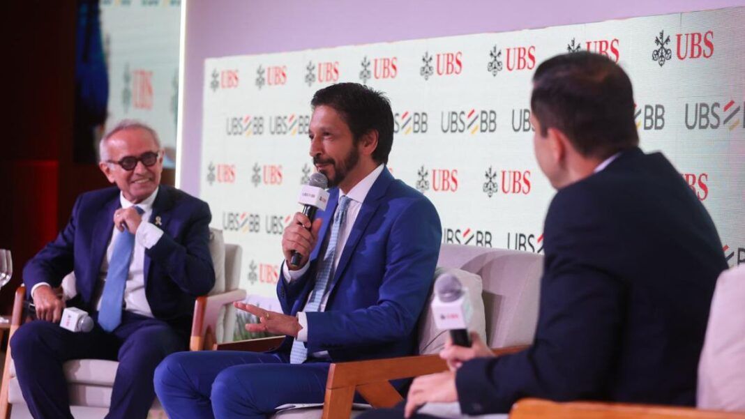 Homens de terno participam de um painel em um evento corporativo, sentados em poltronas e conversando com microfones em mãos. Um painel ao fundo exibe repetidamente os logotipos “UBS” e “UBS BB”. O ambiente é iluminado e formal.