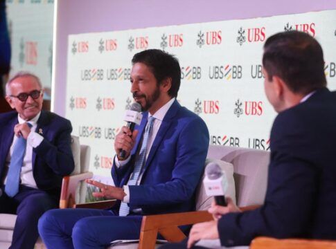 Homens de terno participam de um painel em um evento corporativo, sentados em poltronas e conversando com microfones em mãos. Um painel ao fundo exibe repetidamente os logotipos “UBS” e “UBS BB”. O ambiente é iluminado e formal.