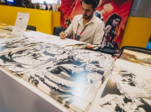 Artista sentado atrás de uma mesa em um evento, desenhando enquanto várias ilustrações em preto e branco de estilo quadrinhos estão expostas à sua frente; ao fundo, painéis coloridos com arte de personagens.