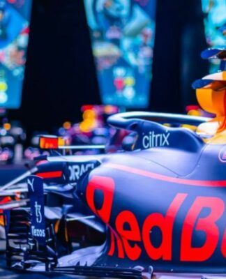 Carro de Fórmula 1 da Red Bull em destaque, com pintura azul e detalhes em vermelho e amarelo, exibido em ambiente iluminado com telas coloridas ao fundo mostrando imagens relacionadas à equipe.