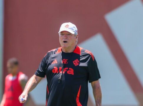 A imagem é um plano médio do técnico de futebol Abel Braga, em pé, em um campo de treinamento durante o dia, sob forte luz solar.