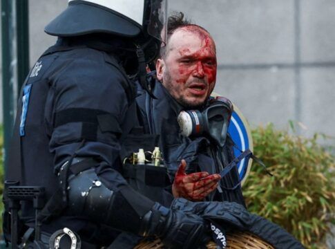 A foto apresenta um plano médio que foca no confronto direto entre um manifestante ferido e um policial da tropa de choque. A cena é dramática e possui um forte impacto visual devido ao contraste entre o sangue e o equipamento policial.