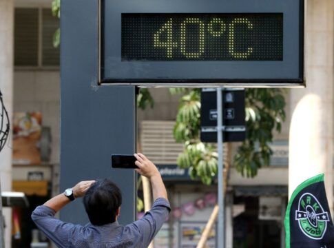 A foto registra um momento típico de calor extremo em um ambiente urbano. O foco central é um termômetro de rua digital que marca uma temperatura de 40ºC, enquanto um pedestre registra a cena.