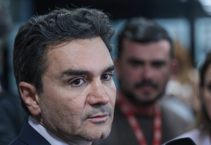 A foto é um primeiro plano (close-up) do rosto de Celso Sabino, atual Ministro do Turismo do Brasil. Ele está posicionado de três quartos, olhando para a direita da imagem com uma expressão séria e atenta, como se estivesse ouvindo uma pergunta ou concedendo uma entrevista.