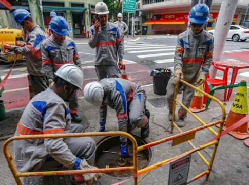 A imagem mostra um grupo de cerca de seis trabalhadores da empresa de energia Enel realizando manutenção ou reparos em uma via urbana de São Paulo. Eles estão em uma pose de trabalho concentrado ao redor de um bueiro ou abertura no chão.