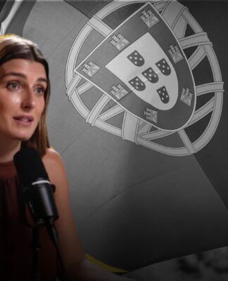 A imagem mostra a jornalista Joana Treptow em um plano médio, sentada e falando em um microfone de estúdio, com uma bandeira de Portugal em preto e branco ao fundo.