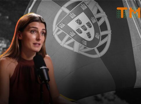 A imagem mostra a jornalista Joana Treptow em um plano médio, sentada e falando em um microfone de estúdio, com uma bandeira de Portugal em preto e branco ao fundo.