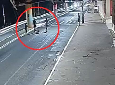 A imagem é um registro de câmera de monitoramento (CFTV), com ângulo alto e diagonal, mostrando uma via pública urbana durante a noite ou madrugada. A iluminação é artificial e fria, vindo de postes de luz. No meio da rua, destacado por um grande círculo vermelho desenhado digitalmente na imagem, há um grupo de três pessoas em pé ao redor de uma quarta figura que parece estar caída ou sentada no chão.