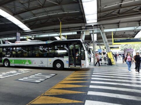 A imagem mostra um terminal de ônibus coberto na cidade de São Paulo, com um veículo de transporte público parado e pessoas circulando na área de embarque e desembarque.