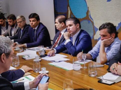 Esta é uma fotografia em ambiente interno que mostra uma reunião de trabalho com a presença de diversos homens, presumivelmente líderes políticos ou assessores, sentados ao redor de uma grande mesa de madeira. O foco principal está nas pessoas sentadas do lado esquerdo da mesa.
