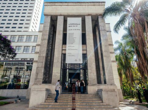 A imagem é uma vista ampla e frontal do edifício principal da Biblioteca Mário de Andrade, mostrando sua arquitetura imponente e o entorno verde, sob luz solar intensa. O prédio é construído em um estilo monumental e clássico, com predomínio de pedra clara ou granito cinza nas fachadas. A entrada principal é marcada por uma grande escadaria que leva a uma plataforma elevada.