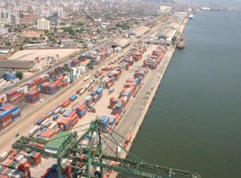 Esta é uma vista aérea ampla que captura uma seção do Porto de Santos no Brasil, destacando a intensa atividade de movimentação de contêineres e a proximidade com a área urbana da cidade.