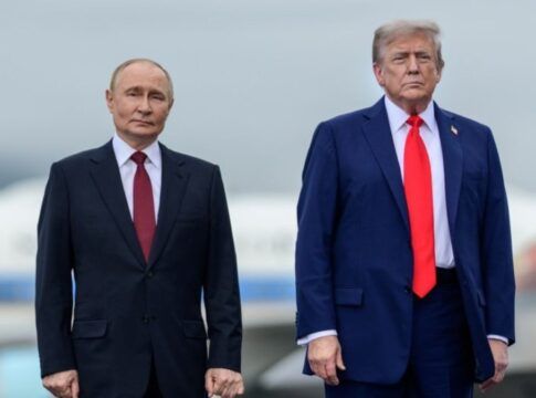 A imagem é um plano médio que captura os presidentes Vladimir Putin e Donald Trump lado a lado, em pé, em um ambiente externo que se assemelha a uma pista de aeroporto ou área externa de recepção, sob um céu nublado.