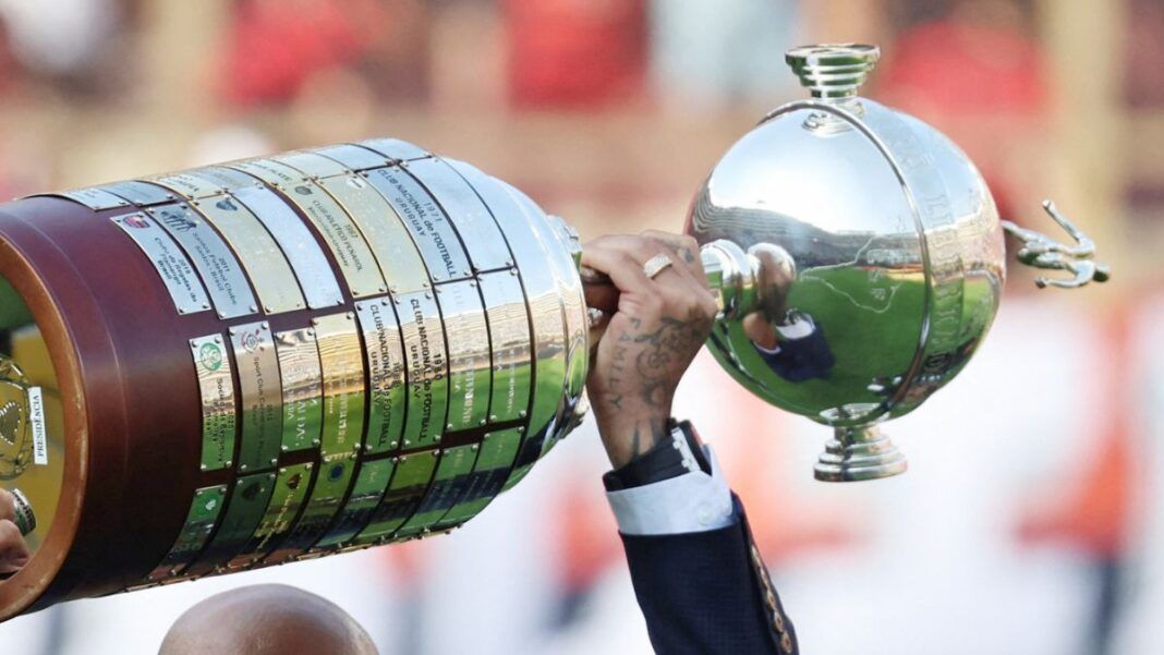 A foto mostra um registro em close-up e em ângulo lateral da Taça Libertadores da América sendo erguida por um jogador. O troféu está na horizontal, ocupando grande parte do quadro.