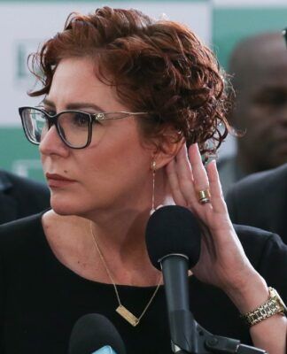 A imagem é um close-up que foca na deputada federal Carla Zambelli em um ambiente formal, possivelmente uma coletiva de imprensa ou evento político, com outras pessoas em segundo plano.