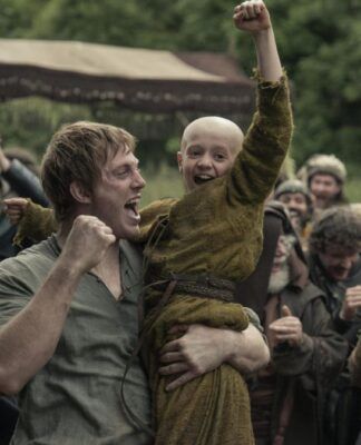 Homem levanta uma criança careca nos braços enquanto ambos comemoram com punhos erguidos; ao redor, um grupo de pessoas vestidas com trajes medievais celebra em um acampamento ao ar livre, com tendas e vegetação ao fundo.