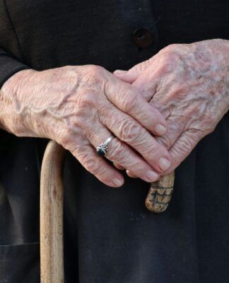 Mãos envelhecidas pelo passar da idade seguram uma bengala de madeira, na mão esquerda, um anel solitário prateado no dedo anelar, aquele que antecede o mindinho.