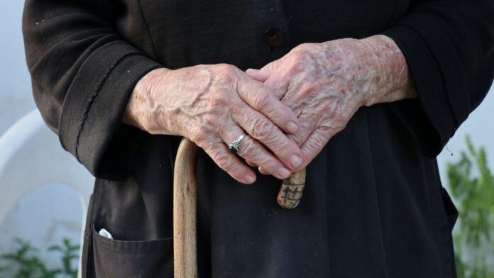 Mãos envelhecidas pelo passar da idade seguram uma bengala de madeira, na mão esquerda, um anel solitário prateado no dedo anelar, aquele que antecede o mindinho.